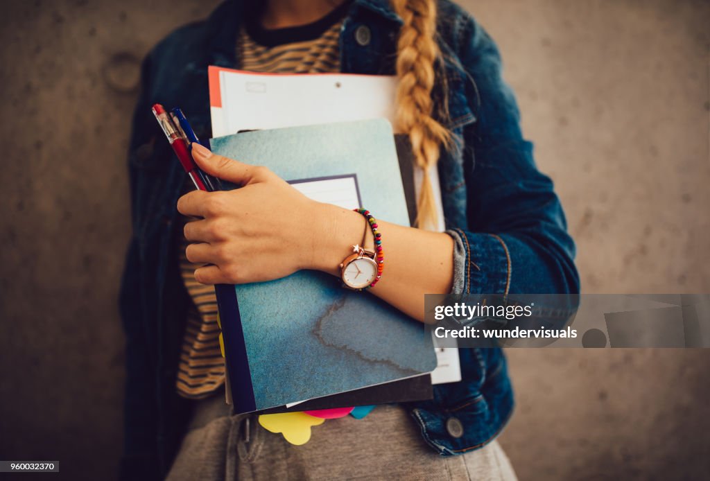 Teenage girl holding books, notebooks and pencils standing against wall