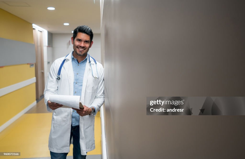 Alegre médico homem encostado na parede segurando uma ficha médica, olhando para a câmera muito feliz