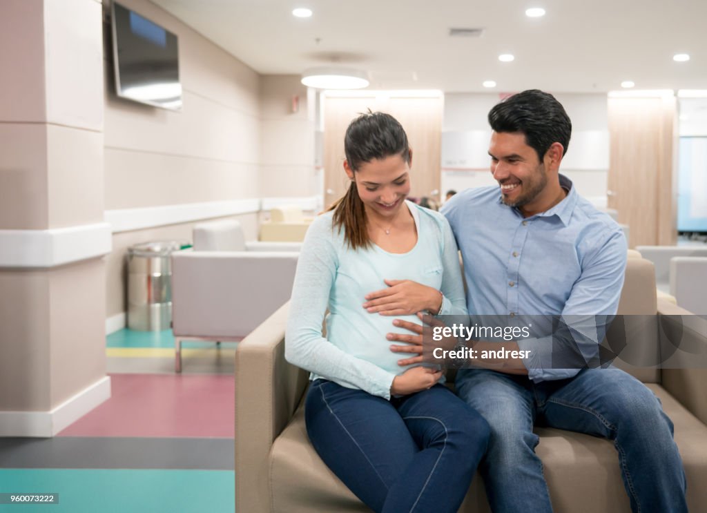 Hermosa pareja jóvenes sentados en la sala de espera del hospital mientras que el marido toca su vientre esperando consulta con obstetra