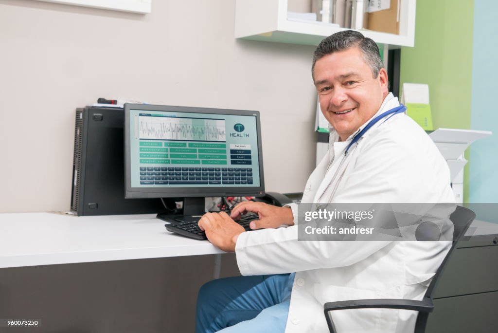 Doctor masculino amistoso completando una historia clínica en ordenador y mirando a cámara muy feliz