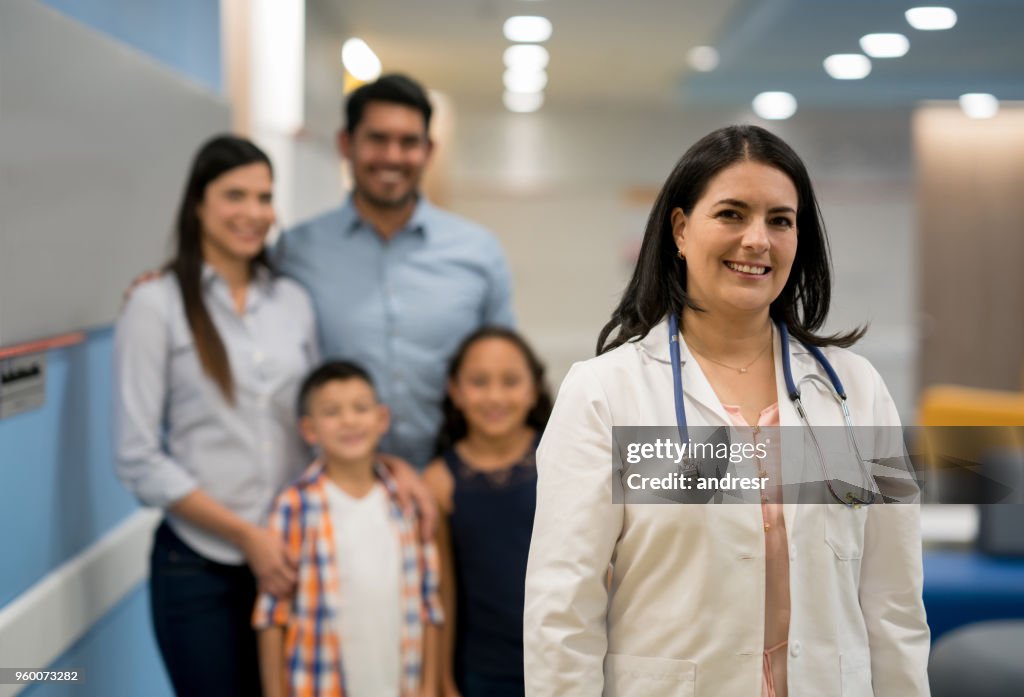 Portait of female mid adult pediatrician looking at camera smiling and a family standing at the background