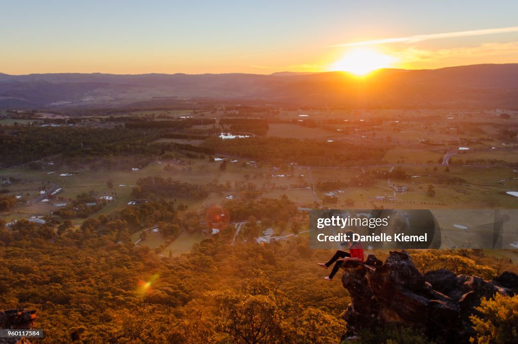 Young woman sitting on cliff taking photo at sunset