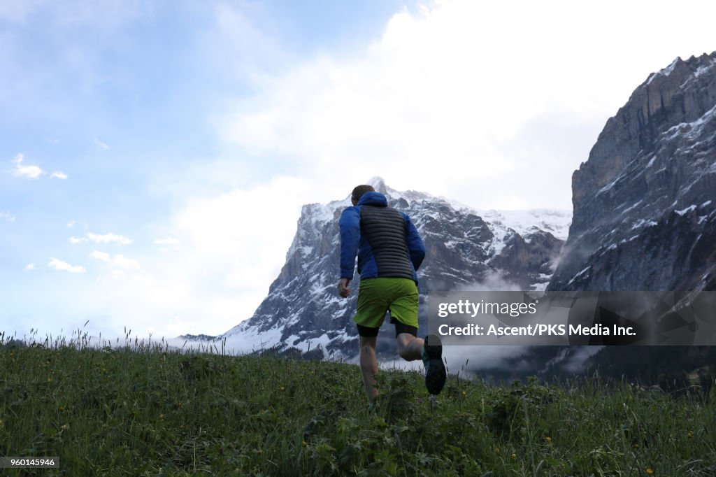 Mountain trail runner traverses meadow, below mountains