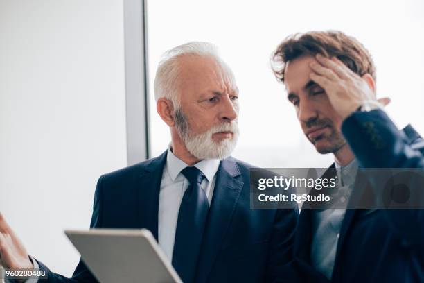 homem de negócios sênior com tablet digital em suas mãos, franzindo a testa, colega de trabalho jovem, segurando a mão na cabeça - franzindo as sobrancelhas - fotografias e filmes do acervo