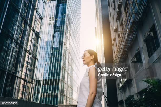 smart young woman surrounded by highrise corporate buildings and looking up into the sky with confidence - über etwas schauen stock-fotos und bilder