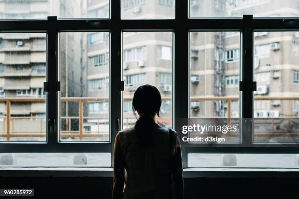 rear view of woman looking out to city through window - a lonely woman stock-fotos und bilder