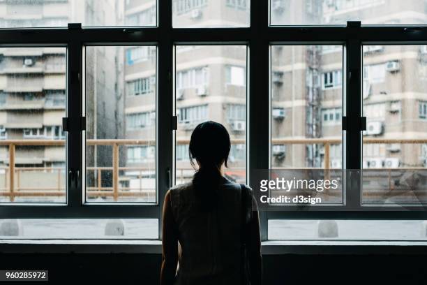rear view of woman looking out to city through window - solitario foto e immagini stock