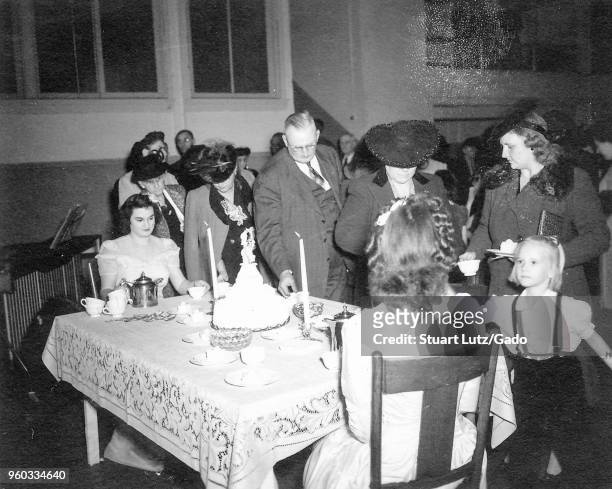 Black and white photograph, showing two women, seated at either side of a small table that is laden with candles, cake, and tea, handing out cups of...