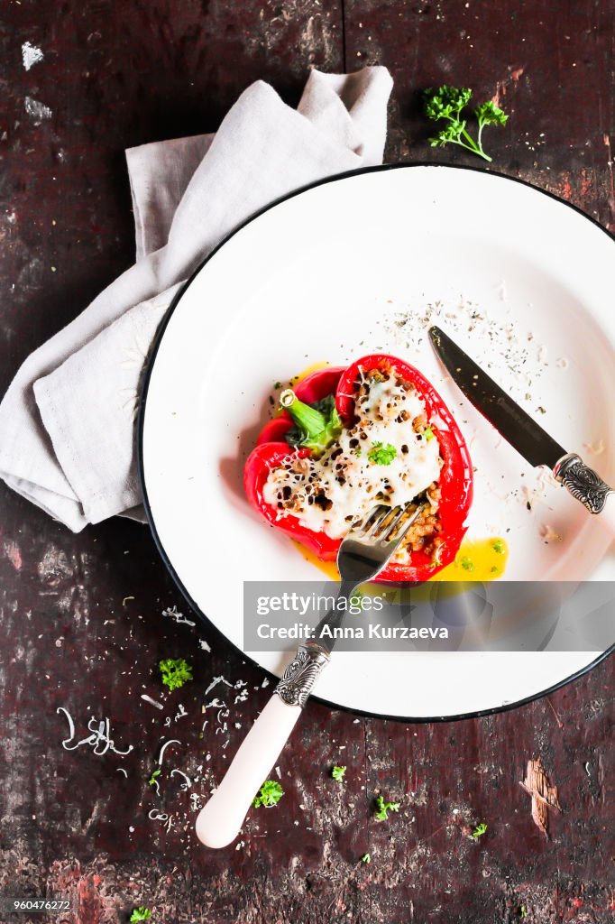 Red bell pepper stuffed with pork and beef meat, carrot, onion with grated cheese sprinkled with spices and fresh parsley in a plate on a wooden table, top view