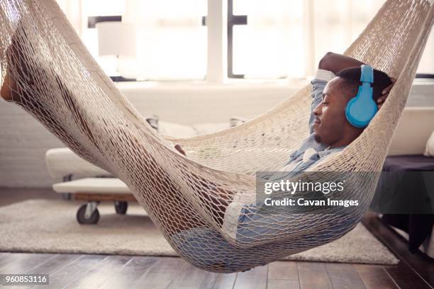 man listening music while resting on hammock in living room at home - young man listening music and relaxing in hammock stock pictures, royalty-free photos & images
