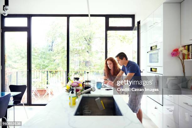 couple using laptop computer while standing at kitchen counter by window - man leaning chair standing side view stock pictures, royalty-free photos & images
