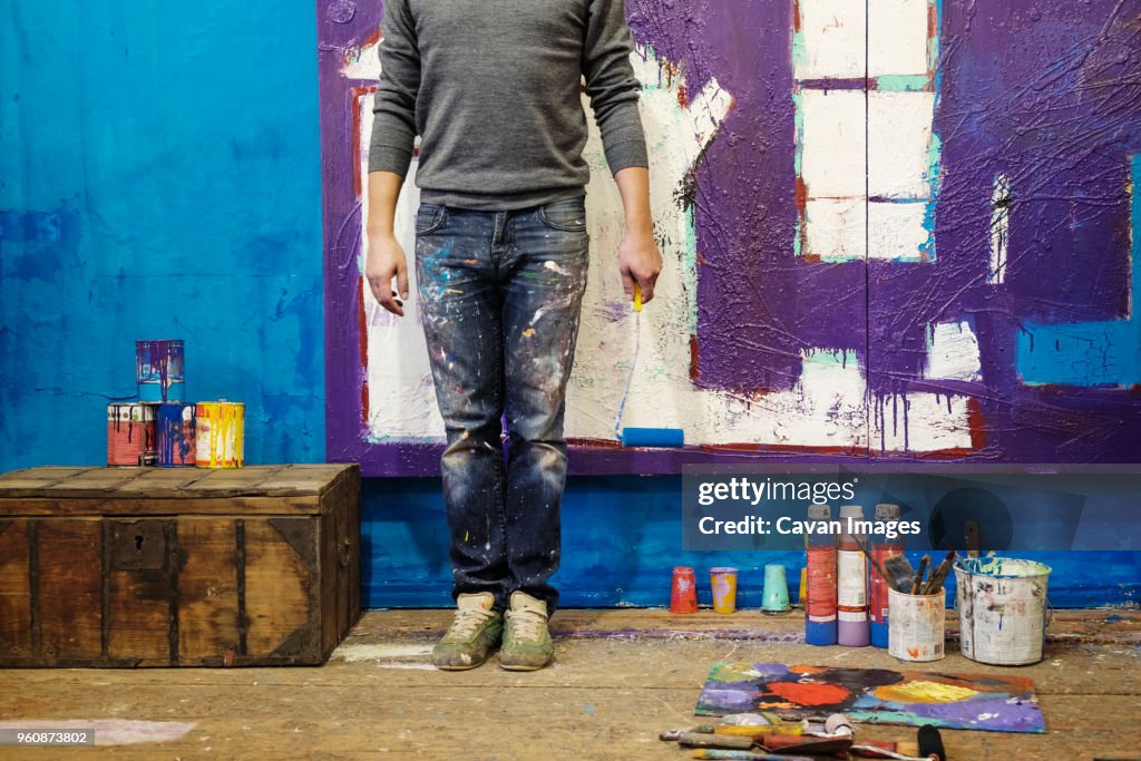Low section of male painter holding paint roller and standing against wall
