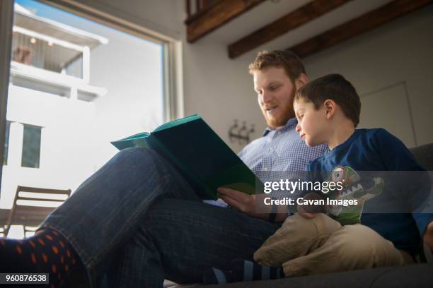 low angle view of father holding book and teaching son on sofa - genderblend2015 stock pictures, royalty-free photos & images