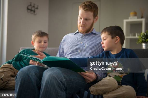 father showing book to sons on sofa at home - genderblend2015 stock pictures, royalty-free photos & images