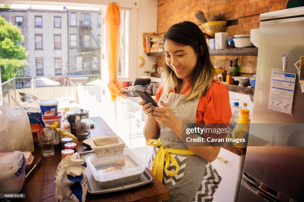 Smiling woman using phone while cooking in kitchen at home
