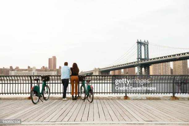 couple standing with bicycles on footpath by manhattan bridge against sky - touristen brooklyn bridge stock-fotos und bilder