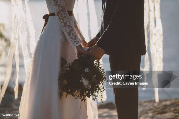 midsection of wedding couple holding bouquet while standing at beach during sunset - sunset beach bouquet wedding couple stock pictures, royalty-free photos & images