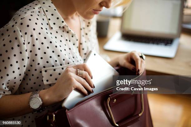 midsection of woman putting tablet computer in purse at home office - ハンドバッグ ストックフォトと画像