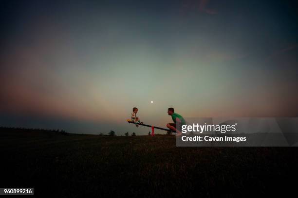 father and son playing on seesaw against sky at dusk - balançoire à bascule photos et images de collection