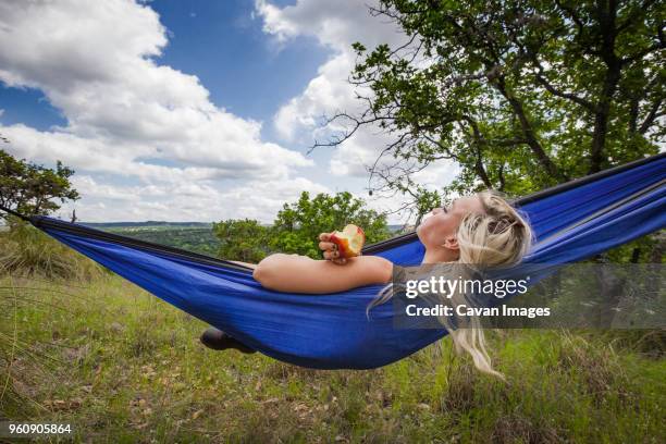 woman holding apple while lying on hammock at field during vacation - hill country stock pictures, royalty-free photos & images