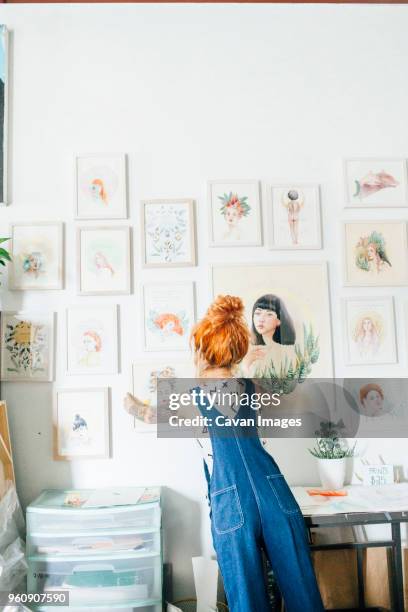 rear view of female artist hanging paintings on wall in studio - artist painting stock-fotos und bilder