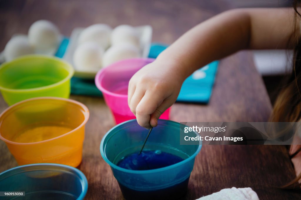 Cropped hand of girl using stirrer to mix dye in water for Easter Egg coloring on table
