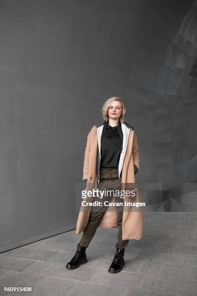 full length portrait of confident woman standing near gray wall - coat stock-fotos und bilder
