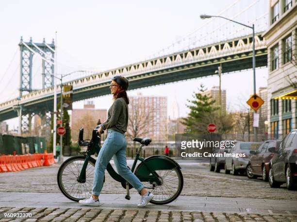 side view of woman with bike share walking on street with manhattan bridge in background - sustainable tourism stock pictures, royalty-free photos & images