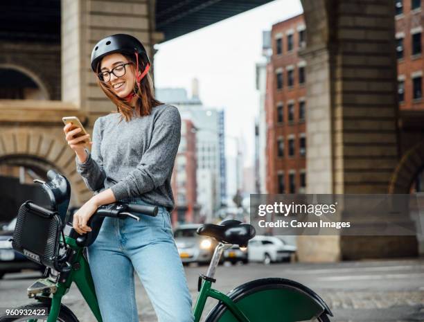 happy woman using smart phone while standing by bicycle on street - bicycle rental stock pictures, royalty-free photos & images