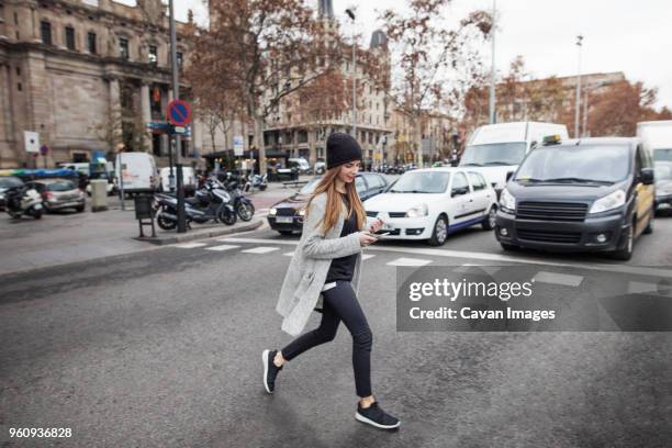 young woman using phone while crossing city street - attraversare foto e immagini stock