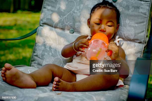baby girl sitting on garden lounge chair drinking from baby cup - african girl drinking water stockfoto's en -beelden