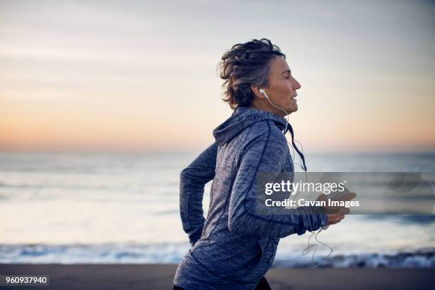woman jogging while listening music at beach against sky - active seniors outdoors stock-fotos und bilder