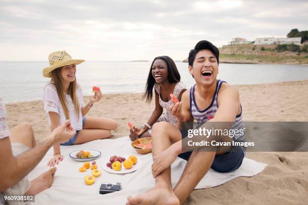 cheerful friends eating fruits while sitting on beach - beach picnic stock pictures, royalty-free photos & images