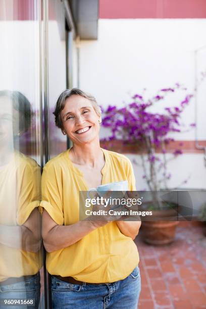 portrait of happy mature woman holding coffee mug while standing at porch - cavan images stockfoto's en -beelden