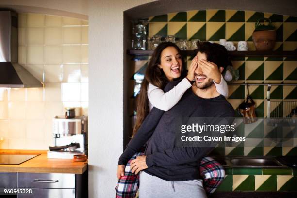 cheerful girlfriend covering boyfriends eyes while standing at home - mani sugli occhi foto e immagini stock