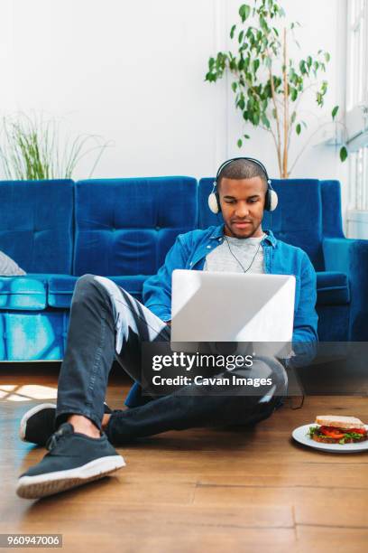 full length of man listening music through laptop computer while sitting on floor in living room - music room stockfoto's en -beelden