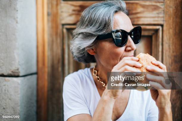 woman in sunglasses eating burger while sitting against door - woman eating burger stockfoto's en -beelden