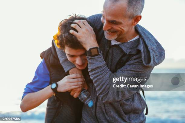 playful father and son playing while exercising at beach against sky - father son going out stock-fotos und bilder