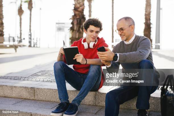 happy father showing smart phone to son while sitting on steps in city - cavan images stock pictures, royalty-free photos & images