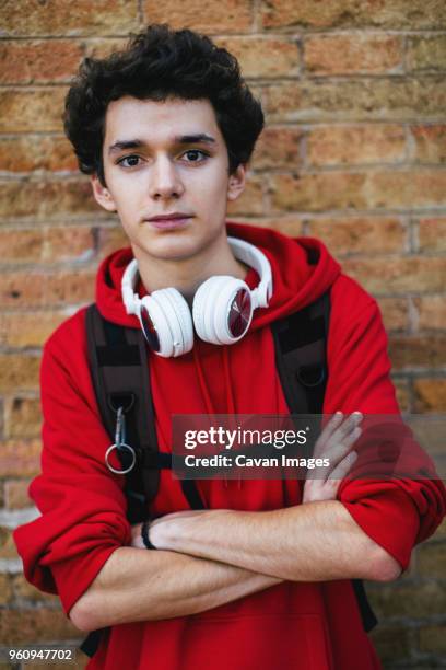 portrait of confident teenage boy with backpack and arms crossed standing against brick wall - one teenage boy only fotografías e imágenes de stock