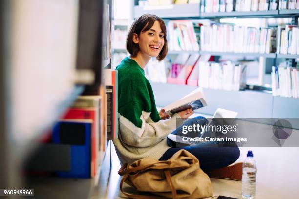 portrait of woman with laptop computer reading book while sitting on floor in library - student reading book stockfoto's en -beelden