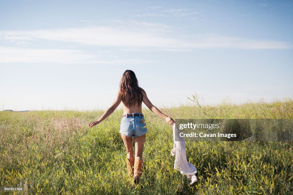 Rear view of woman holding shirt and walking on grassy field against sky