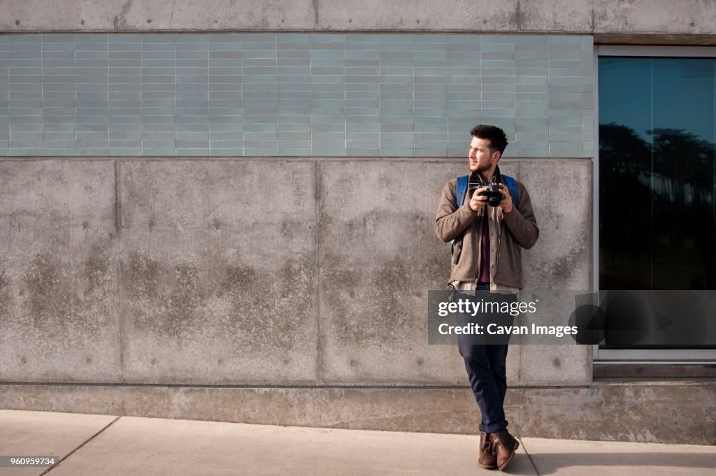 Full length of thoughtful male tourist holding camera while standing on sidewalk