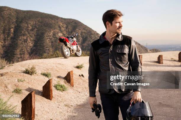 thoughtful motorcyclist holding helmet while standing on mountain - sturzhelm stock-fotos und bilder