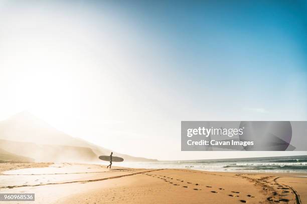 man with surfboard walking towards sea at beach against sky during foggy weather - fuerteventura stock-fotos und bilder