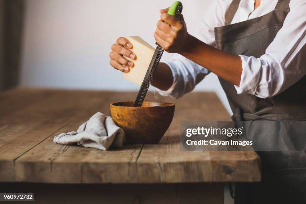 midsection of woman grating cheese in bowl while standing by wooden table - grater stock pictures, royalty-free photos & images