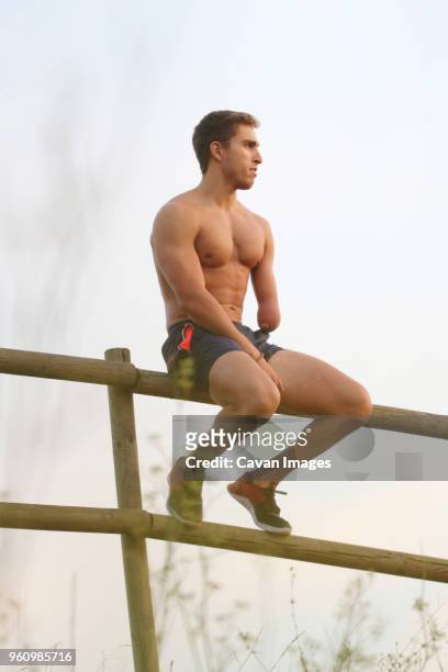 low angle view of thoughtful physically challenged shirtless man looking away while sitting on fence against clear sky - tronco nu imagens e fotografias de stock