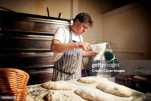 male baker sprinkling flour on bread dough at bakery - cake shop stock pictures, royalty-free photos & images