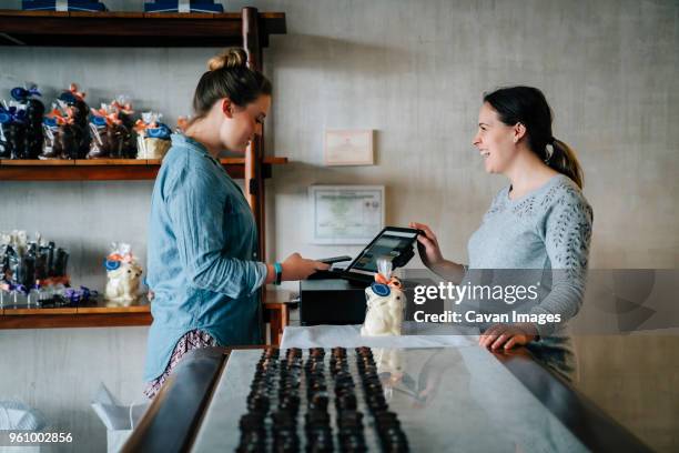 side view of owner laughing while female customer making mobile payment at store - fábrica-de-chocolate fotografías e imágenes de stock