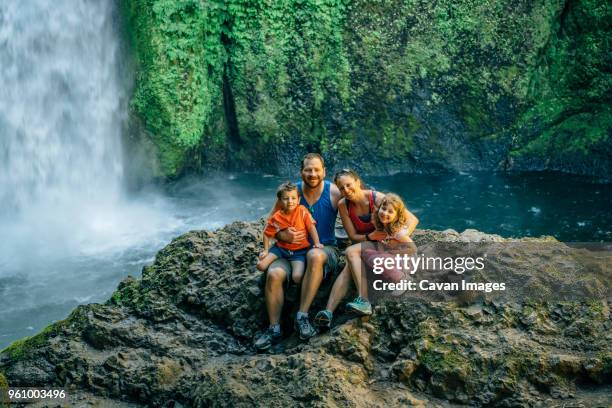 portrait of happy family sitting on rocks against waterfall at forest - front view portrait of four children sitting on rock stock pictures, royalty-free photos & images